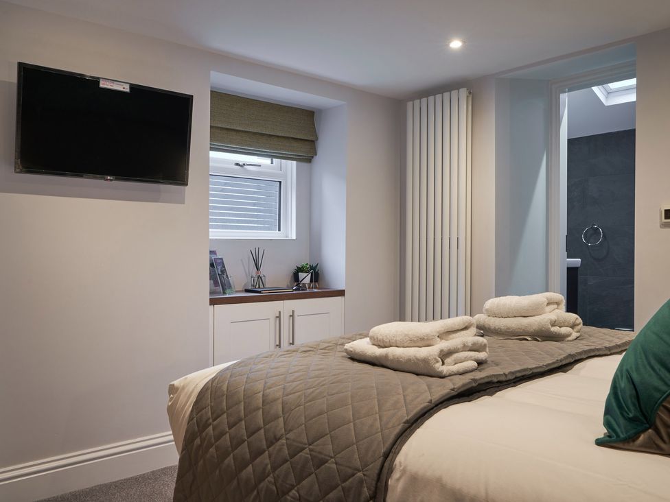 A bedroom with a bed covered with a quilt and folded towels with a flat screen television on the wall a window with a blind and a door leading to a bathroom at Lower Sheriff's Place in Windermere