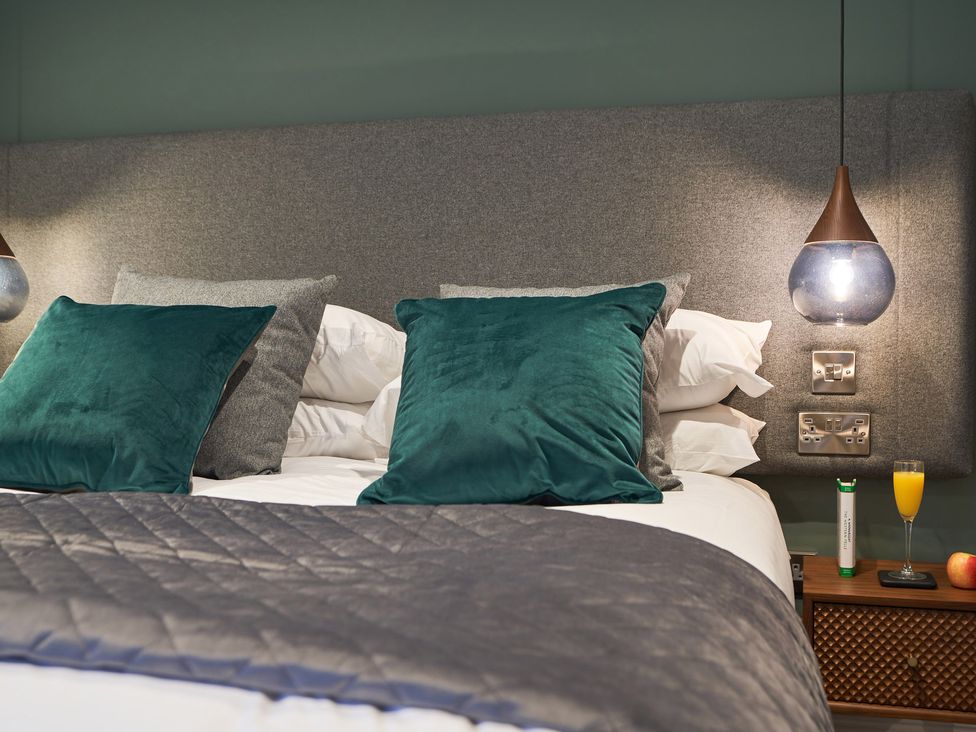 A bed with green and gray pillows a gray quilted blanket a bedside table with a glass of orange juice an apple and a tube at Lower Sheriff's Place in Windermere