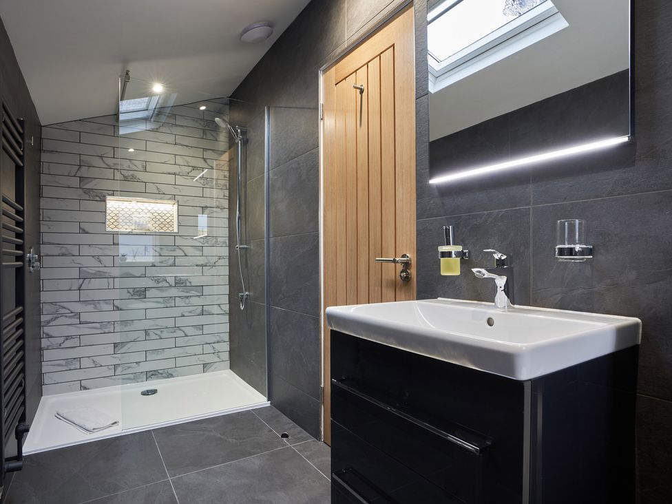 A bathroom with a walk-in shower with white marble tiles a wooden door a black vanity with a white sink and a backlit mirror at Lower Sheriff's Place in Windermere