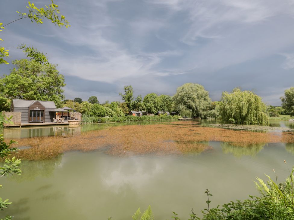 A scenic view of a lake with trees and a cabin at Oak Lodge At Bridge Lake Farm & Fishery Chacombe