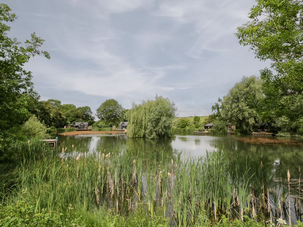 A pond with grass and trees with houses in the background at Oak Lodge At Bridge Lake Farm & Fishery Chacombe