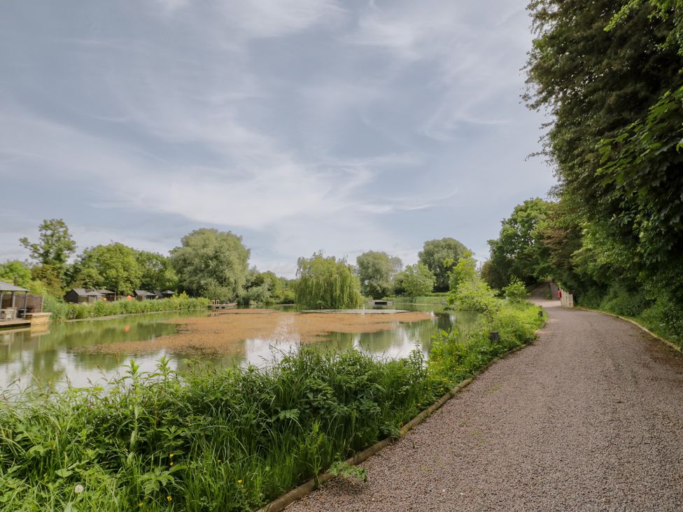 A path next to a pond with greenery surrounding at Oak Lodge At Bridge Lake Farm & Fishery Chacombe