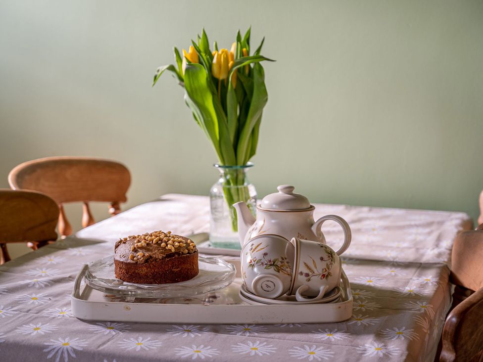 A dining room table with a cake, teapot, teacup, and flowers at Marmaduke Cottage in Market Weighton