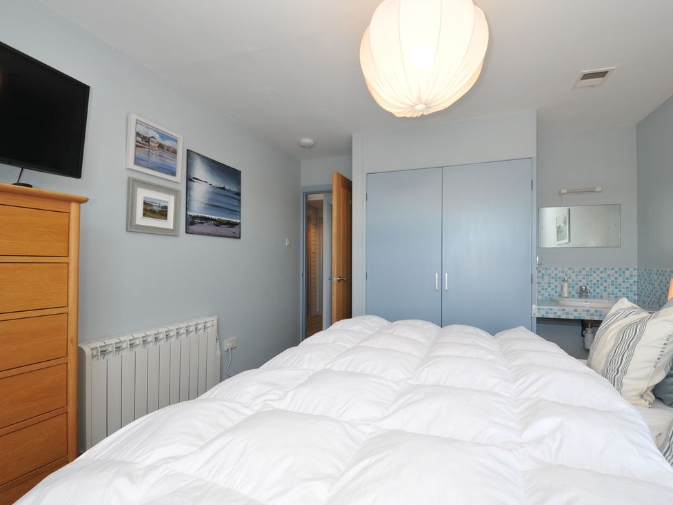 A bedroom with a bed covered in white bedding a wooden dresser television on the wall blue wardrobe doors a small sink with a mirror and paintings on the wall at Slipway 16 in Rock