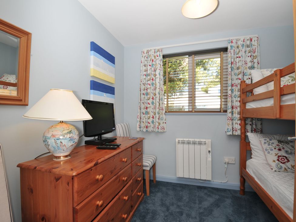 A bedroom with wooden bunk beds a wooden dresser with a lamp and a television a window with floral curtains and a chair at Slipway 16 in Rock