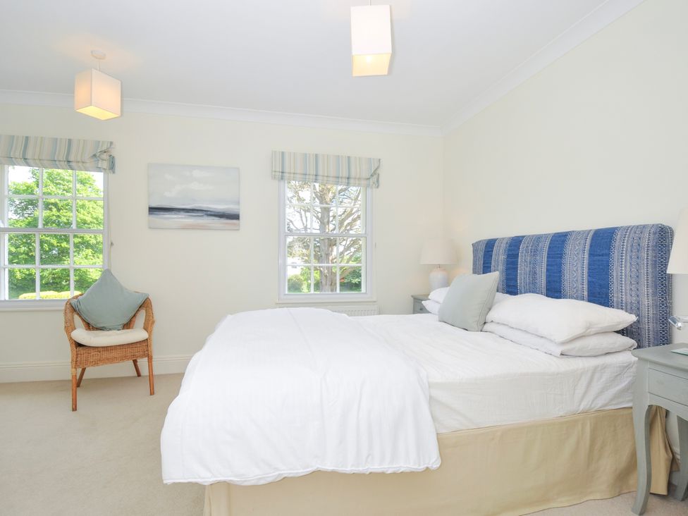 A bedroom with a bed with blue patterned headboard two windows a wicker chair and bedside tables at Lowenna Manor 3 in Rock
