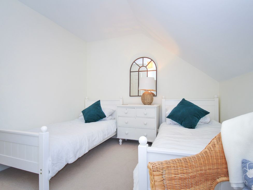 A bedroom with two single beds a white chest of drawers with a lamp and mirror and a wicker chair at Lowenna Manor 3 Rock