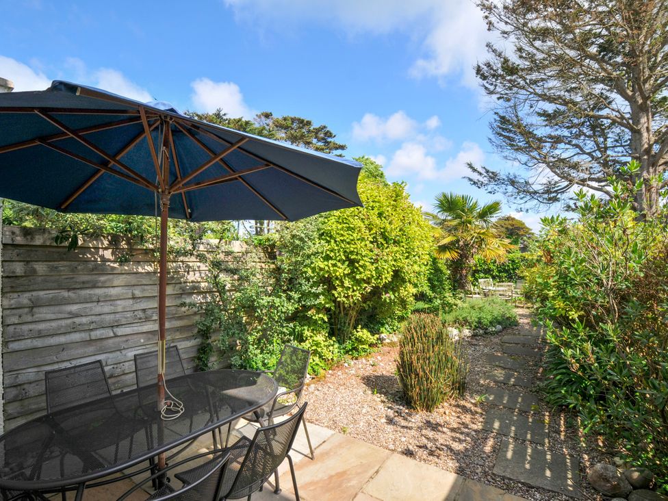 An outdoor patio area with a black metal table and chairs under a large blue umbrella next to a wooden fence and a garden path surrounded by various plants and trees at Lowenna Manor 3 Rock