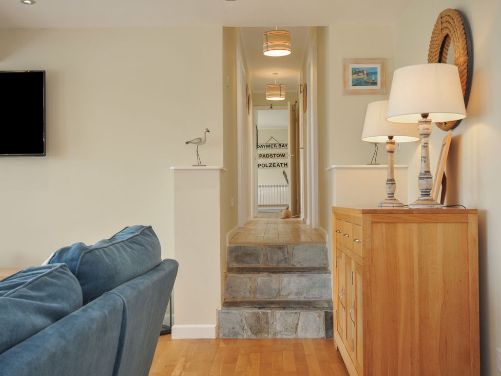 A hallway with stone steps leading to a corridor with ceiling lamps and a wooden cabinet with lamps beside a blue sofa at Dolphins in Daymer Bay