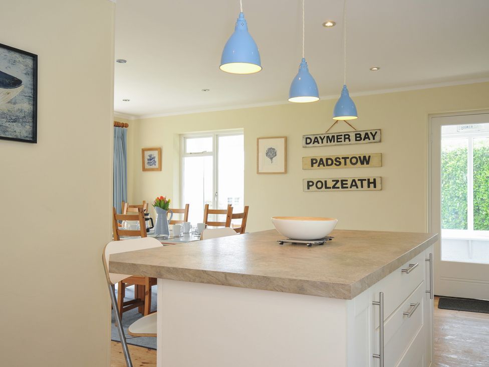 A kitchen island with a bowl under blue pendant lights and a dining area with wooden chairs and a table set with cups and a vase of flowers at Dolphins in Daymer Bay