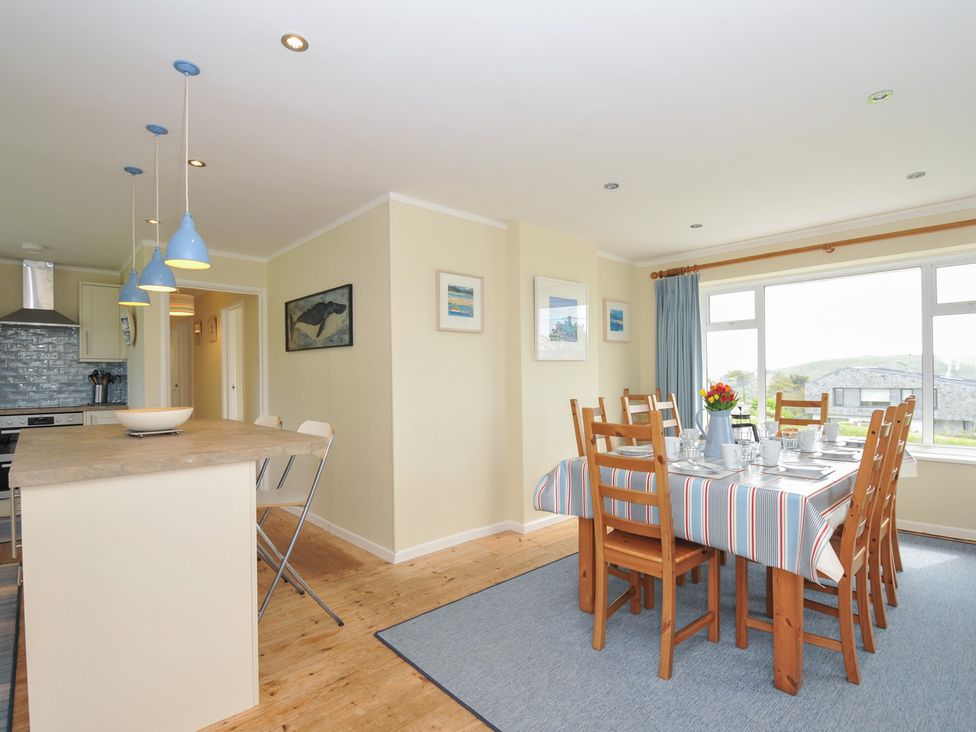 A dining area with wooden chairs and a table set with a striped tablecloth and a kitchen island with blue pendant lights at Dolphins in Daymer Bay