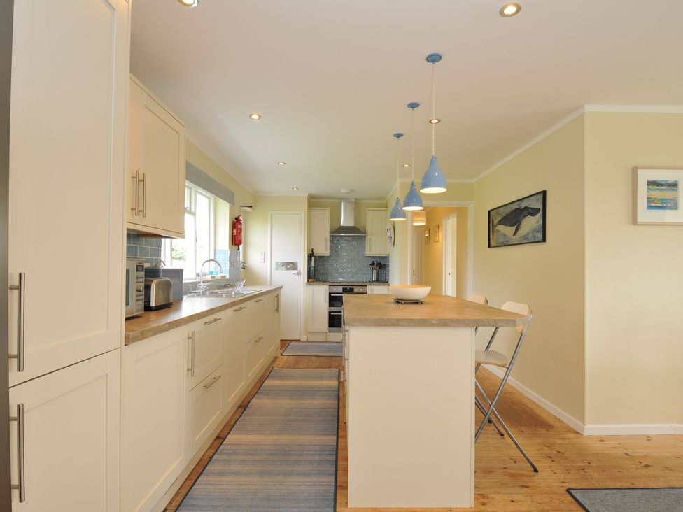 A kitchen with white cabinets a wooden island with chairs and blue pendant lights at Dolphins in Daymer Bay
