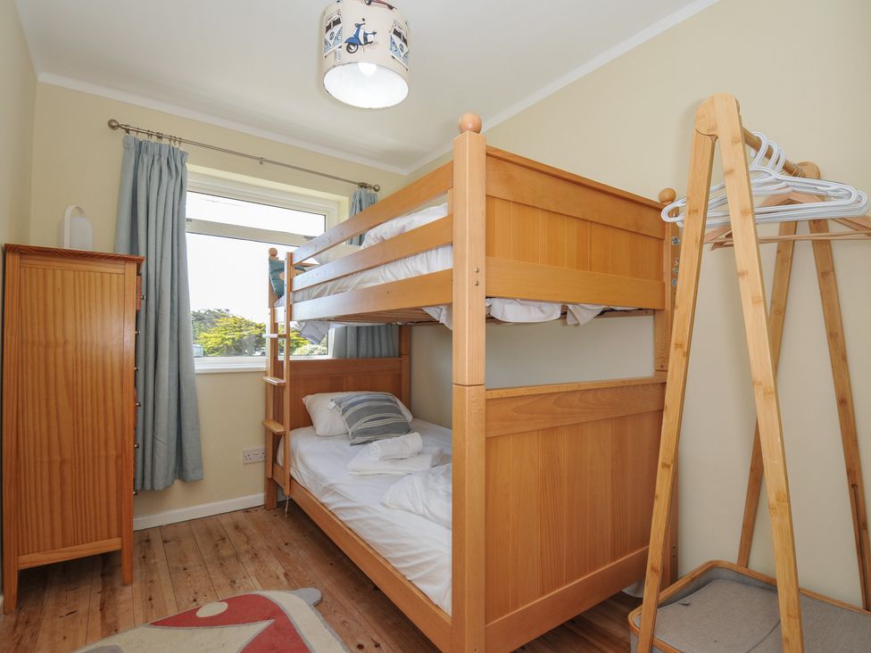 A bedroom with a wooden bunk bed a wooden chest of drawers a wooden clothes rack with hangers and a window with blue curtains at Dolphins in Daymer Bay