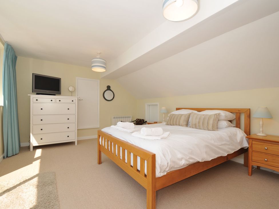 A bedroom with a wooden bed with white linens two bedside tables with lamps a white dresser with a television and a round mirror on the wall at Dolphins Daymer Bay