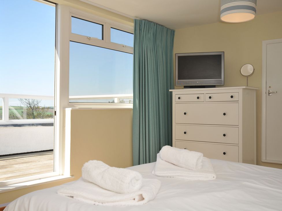 A bedroom with a bed, white towels on the bed, a white chest of drawers with a television on top and a window with blue curtains at Dolphins in Daymer Bay