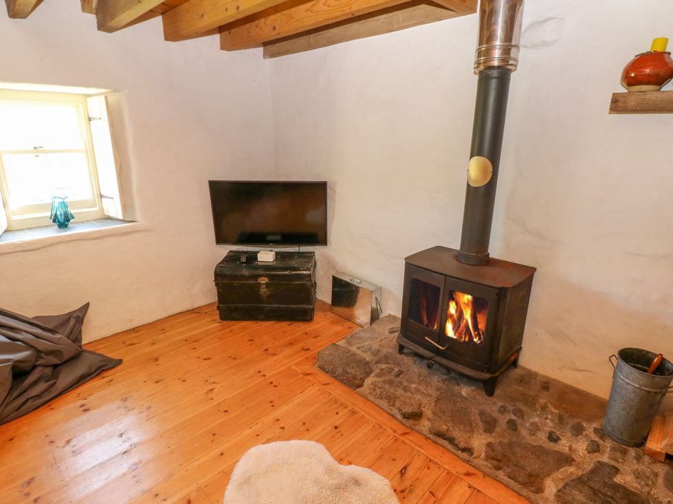 A room with a wood burning stove on a stone hearth a television on a black chest a window and wooden beams on the ceiling at Ty Mellen in Dinas Cross