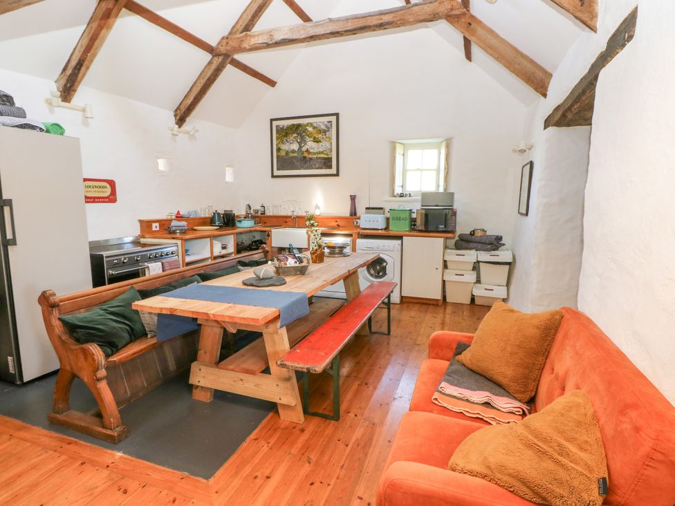 A kitchen with wooden table and bench seating a red sofa with cushions and kitchen appliances at Ty Mellen in Dinas Cross