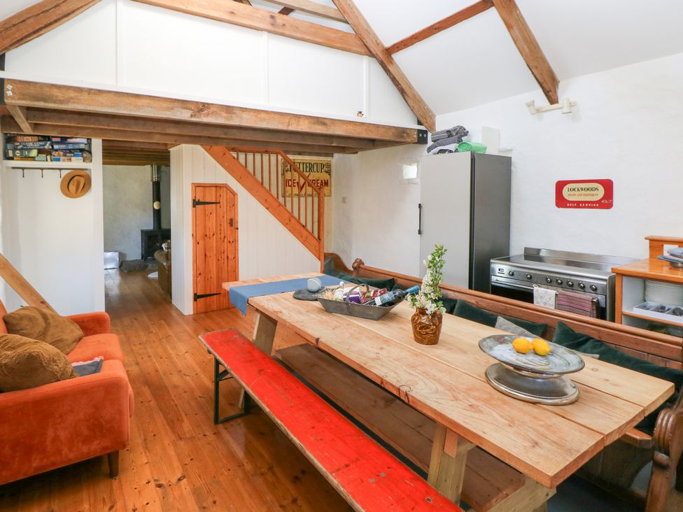 A dining area with a wooden table and red bench adjacent to a kitchen space with a fridge and stove at Ty Mellen in Dinas Cross