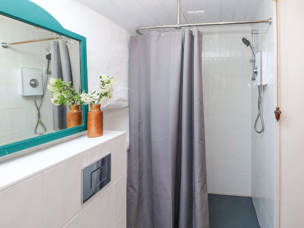 A bathroom with a shower area with a gray shower curtain a showerhead and a white tiled wall a mirror with a green frame and a vase with white flowers on a tiled counter at Ty Mellen in Dinas Cross