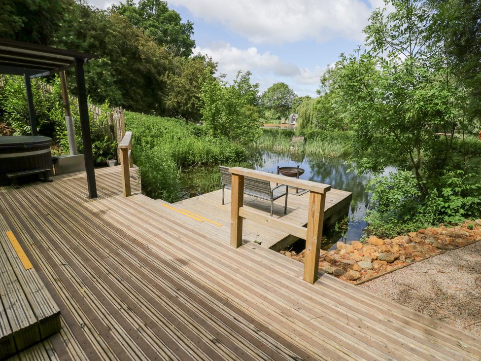 A deck overlooking a pond with seating at The Shepherds Hut at Bridge Lake Farm & Fishery Chacombe