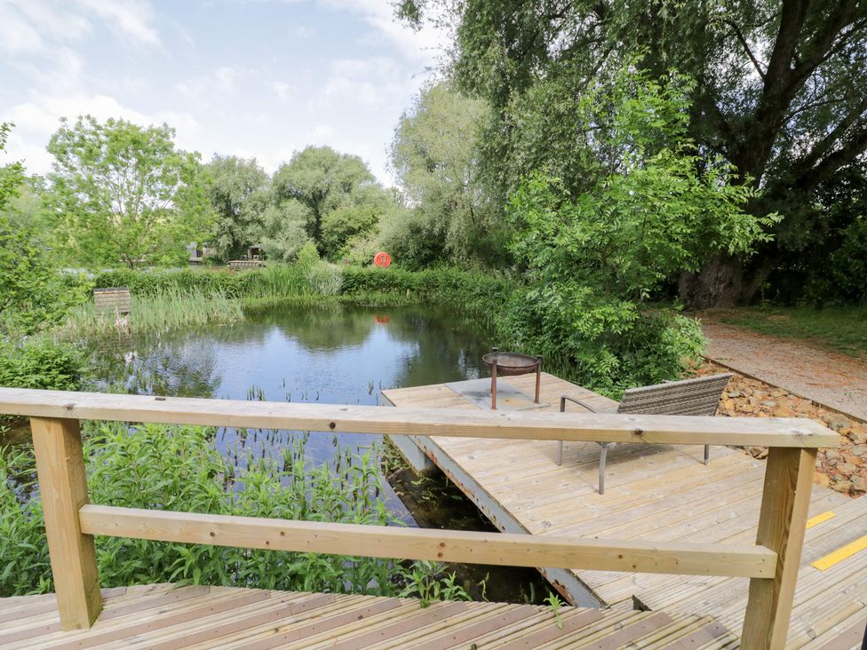 A pond with a wooden deck and a chair at The Shepherds Hut at Bridge Lake Farm & Fishery Chacombe