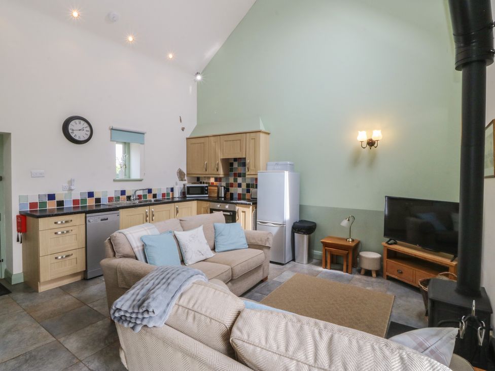 A living room with a kitchen area featuring a clock and a sofa at No 1 The Granary in Capel Coch near Llannerch-Y-Medd
