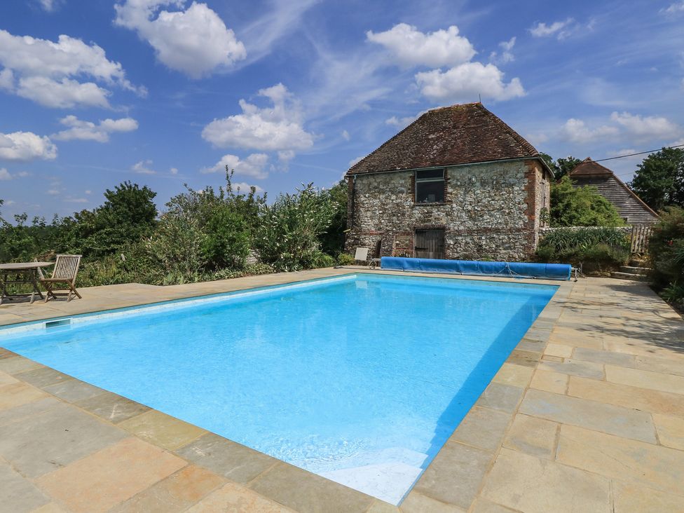 A swimming pool with a stone patio and a house in the background at Byre Cottage 2 Sullington near Storrington