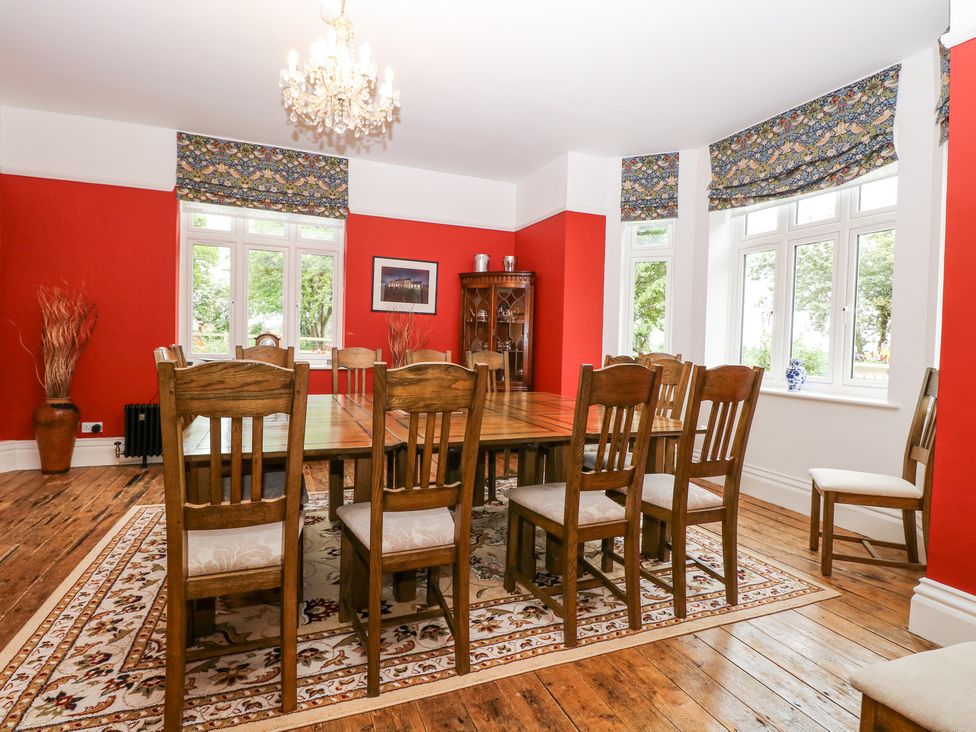 A dining room with a wooden table and chairs on a patterned rug with red walls and windows at High House in Southrepps