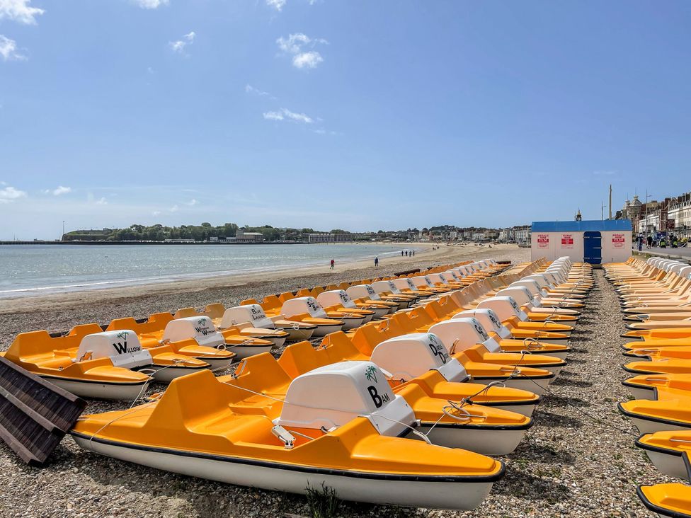 A beach with rows of yellow paddle boats lined up near the shore by the sea with buildings in the background at Corvesgate in Nottington near Weymouth