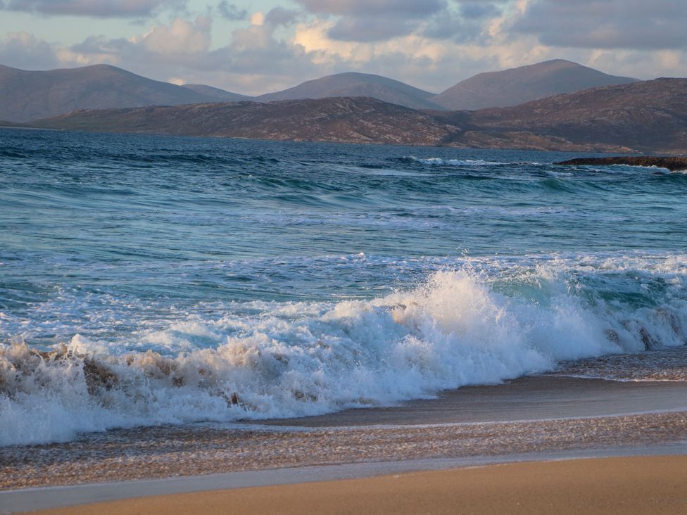 An ocean with waves and sandy beach with mountains in the background at Corncrake Cottage in Leverburgh