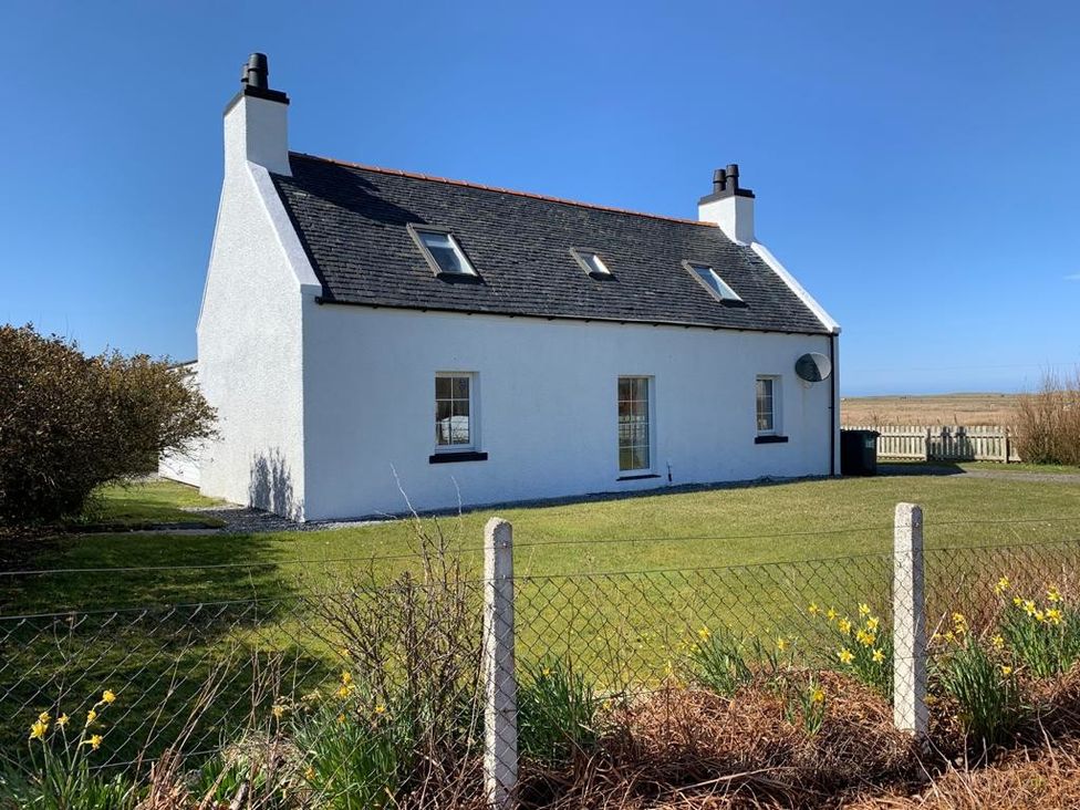 A white cottage with a dark roof and chimneys surrounded by a garden with flowers and grassy lawn at Corncrake Cottage (SU) in Daliburgh