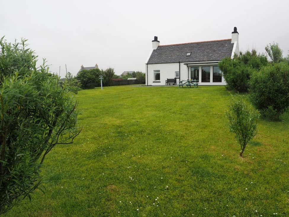A house with two chimneys and a large lawn with bushes and a picnic table at Corncrake Cottage SU in Daliburgh