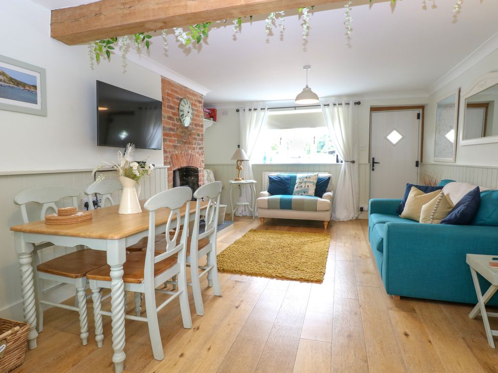 A living room with a dining table and chairs a teal sofa and a beige armchair by a window at Herbies Cottage in Snettisham