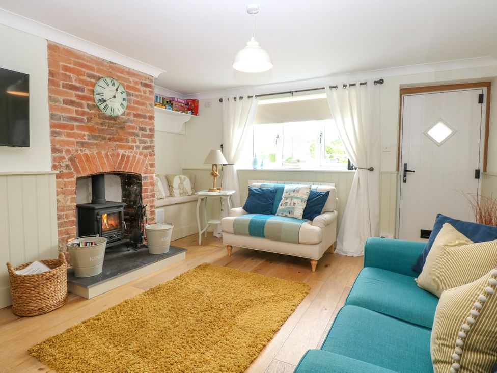 A living room with a brick fireplace wood stove beige armchair teal sofa yellow rug and a white door with window at Herbies Cottage in Snettisham