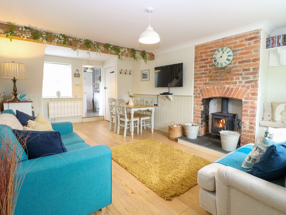 A living room with two sofas a yellow rug a wooden dining table with chairs a brick fireplace with a clock and a mounted television at Herbies Cottage in Snettisham