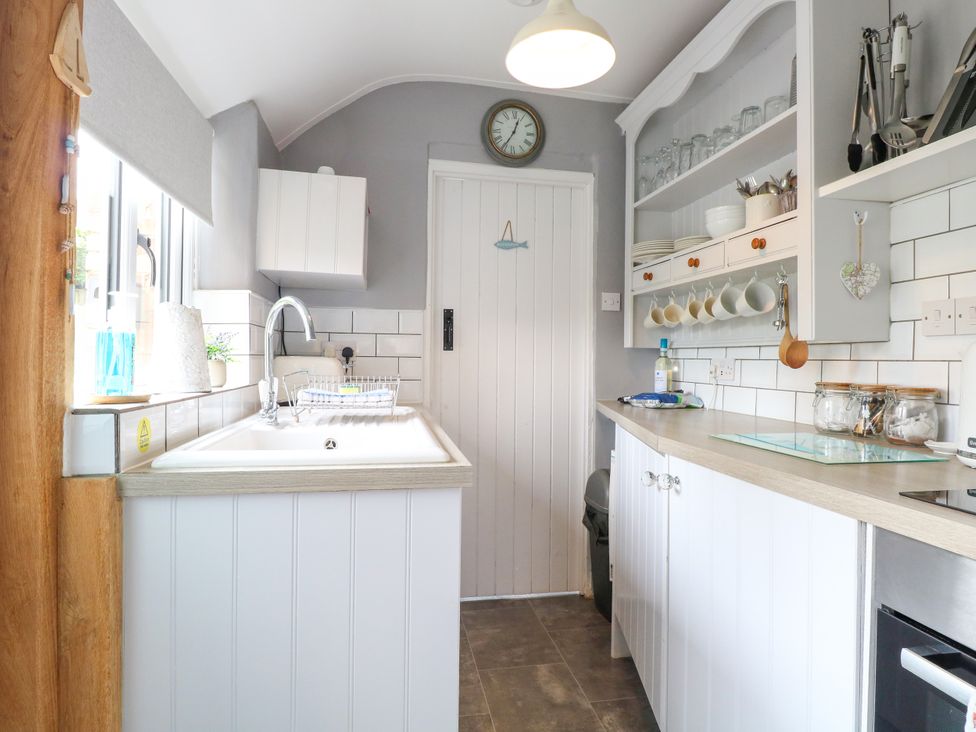 A narrow kitchen with a sink, countertop, hanging cups, shelves with dishes, and a clock above a door at Herbies Cottage in Snettisham