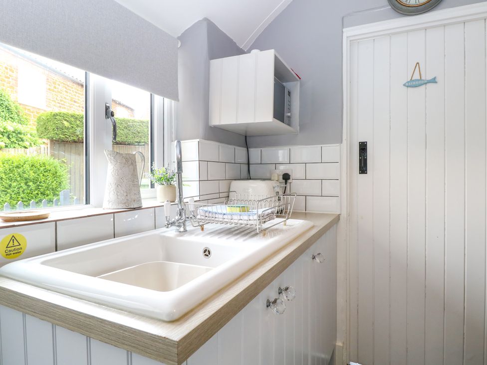 A kitchen sink with a dish rack and a faucet under a window with a pitcher and a plant on the sill and a microwave mounted on the wall next to a white door with a fish-shaped bathroom sign at Herbies Cottage in Snettisham