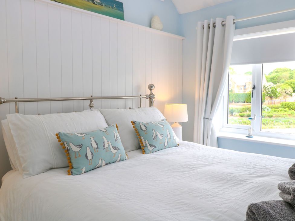 A bedroom with a bed with white bedding and pillows with bird designs beside a window with white curtains and a lamp on a bedside table at Herbies Cottage in Snettisham