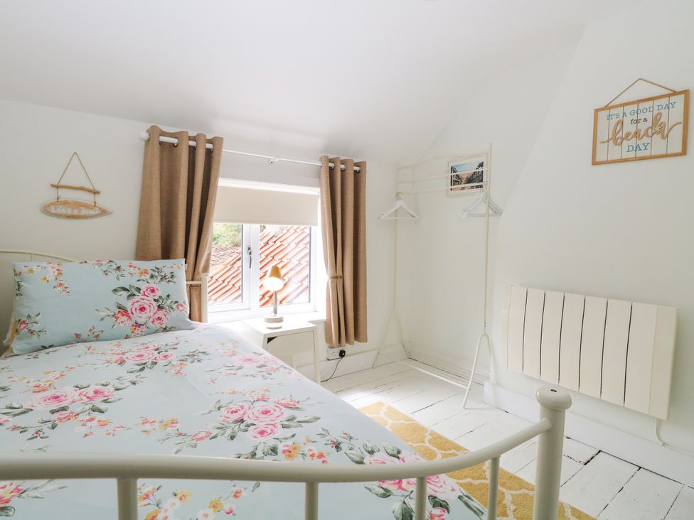 A bedroom with a floral bedspread a window with brown curtains a small lamp a white radiator and a clothes rack at Herbies Cottage in Snettisham