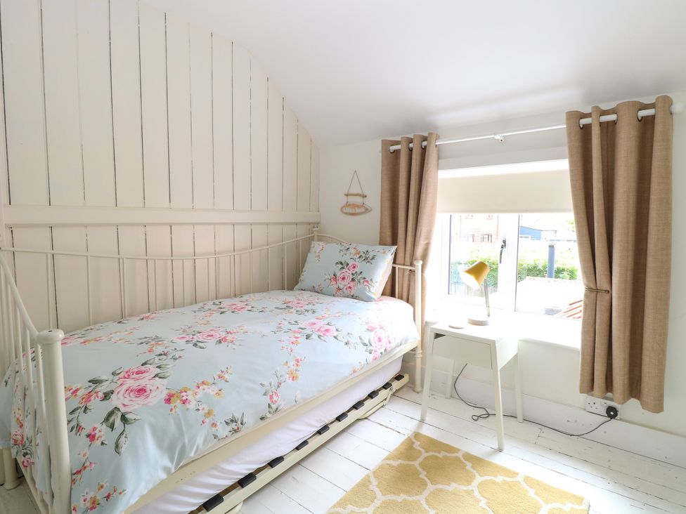 A small bedroom with a single bed with floral bedding a window with brown curtains and a small white desk with a lamp at Herbies Cottage in Snettisham