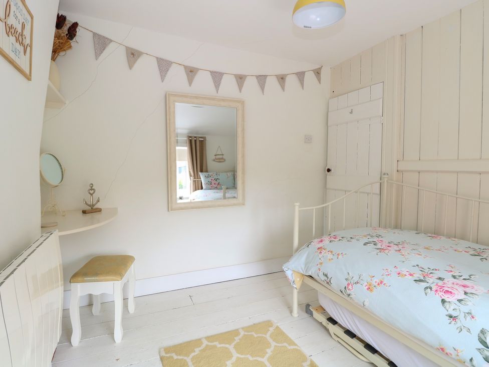 A bedroom with a single bed with floral bedding a small cushioned stool a wall mirror bunting on the wall and a yellow rug at Herbies Cottage in Snettisham
