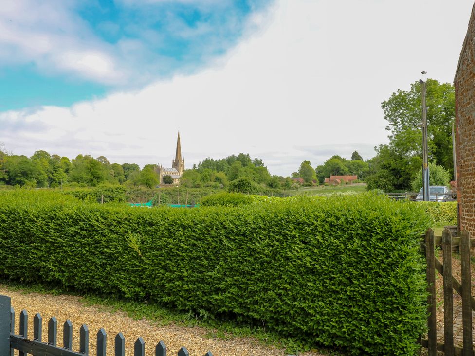A garden with a dense green hedge and a gravel path with a church steeple and trees in the background at Herbies Cottage in Snettisham