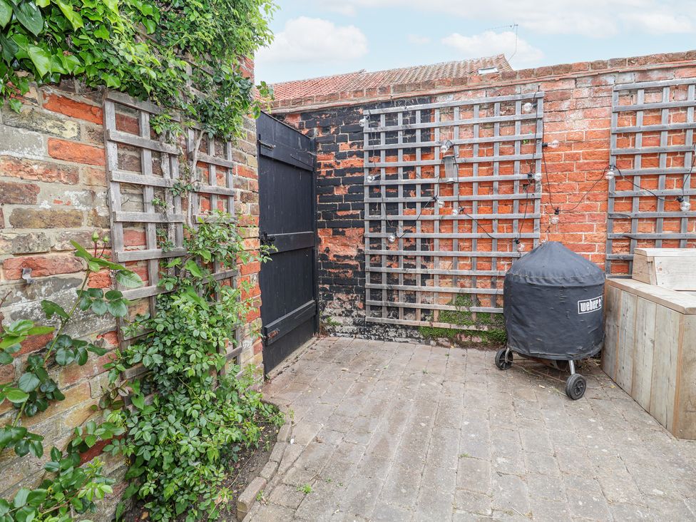 A brick patio with lattice panels on walls a covered barbecue grill and wooden seating at Dolphin Cottage Southwold