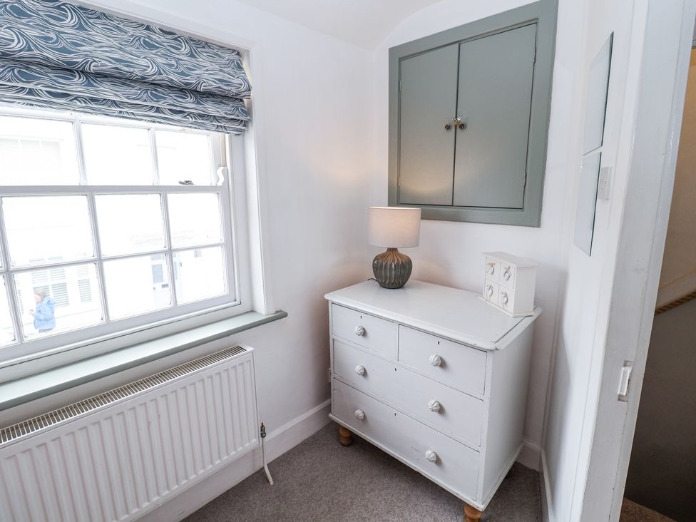 A corner of a room with a white chest of drawers lamp and small cabinet under a closed wall cupboard at Dolphin Cottage Southwold