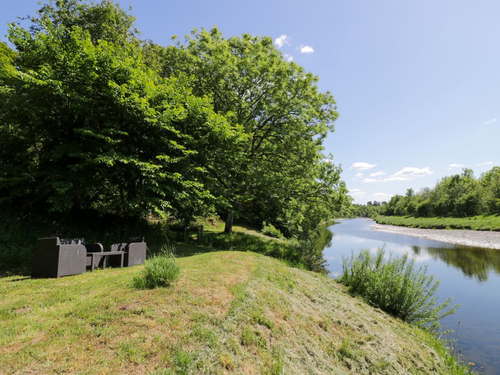 A riverside seating area with trees and grass at Nithbank Cottage