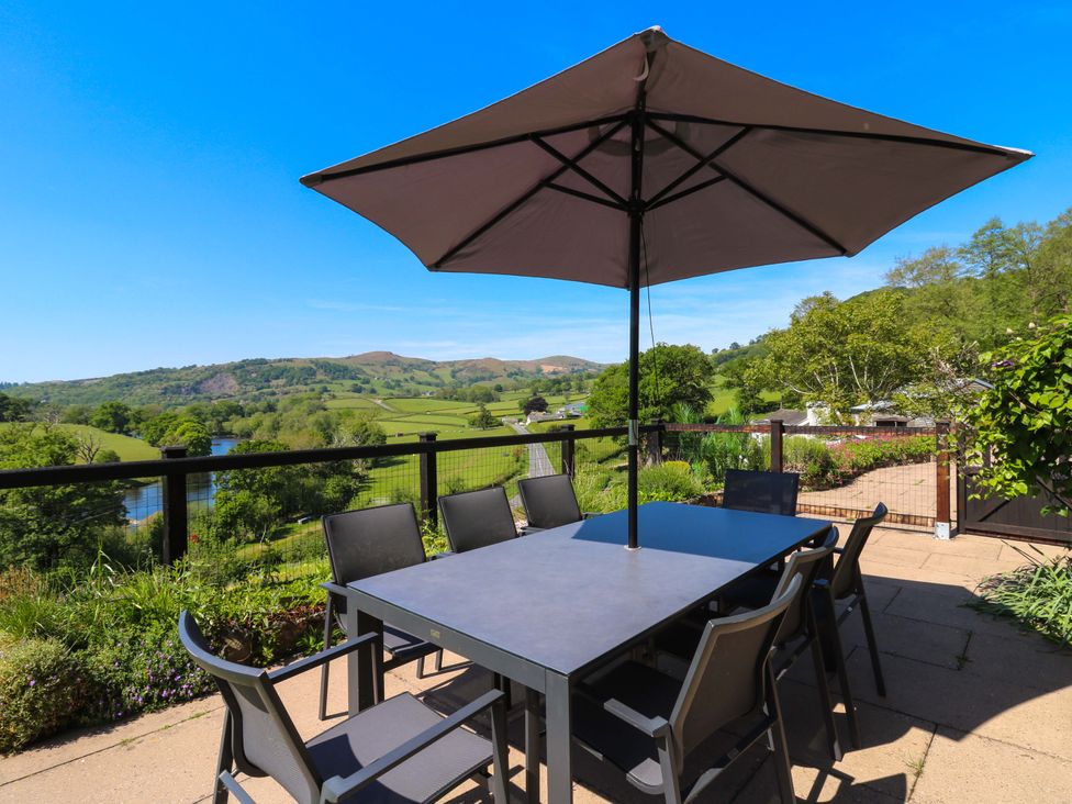 An outdoor patio with a table, chairs, and an umbrella overlooking green hills and a river at Pen Y Banc in Llanfaredd near Builth Wells
