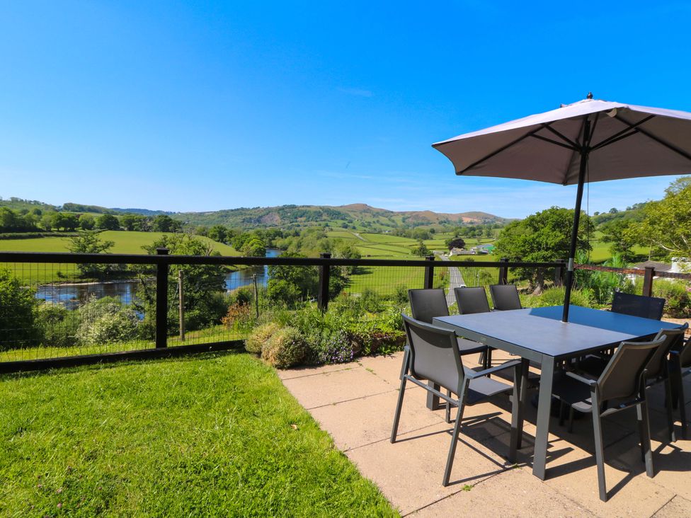 A patio with a table and chairs under an umbrella overlooking a river and green hills at Pen Y Banc in Llanfaredd near Builth Wells