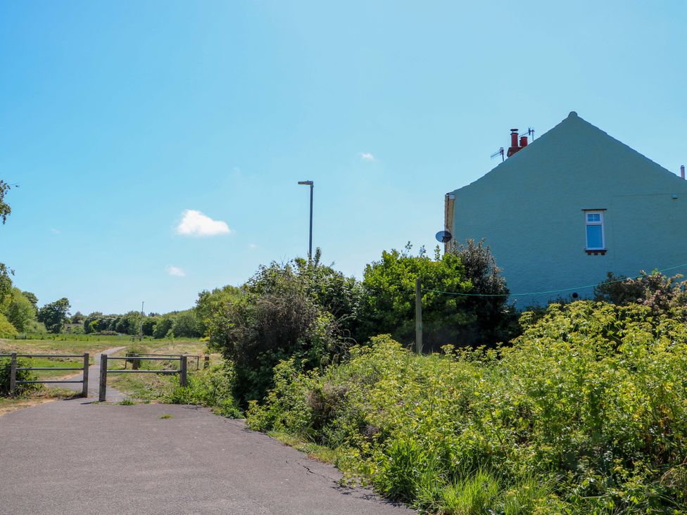A paved path with a wooden gate next to a house and bushes under a blue sky at Beck View in Sheringham