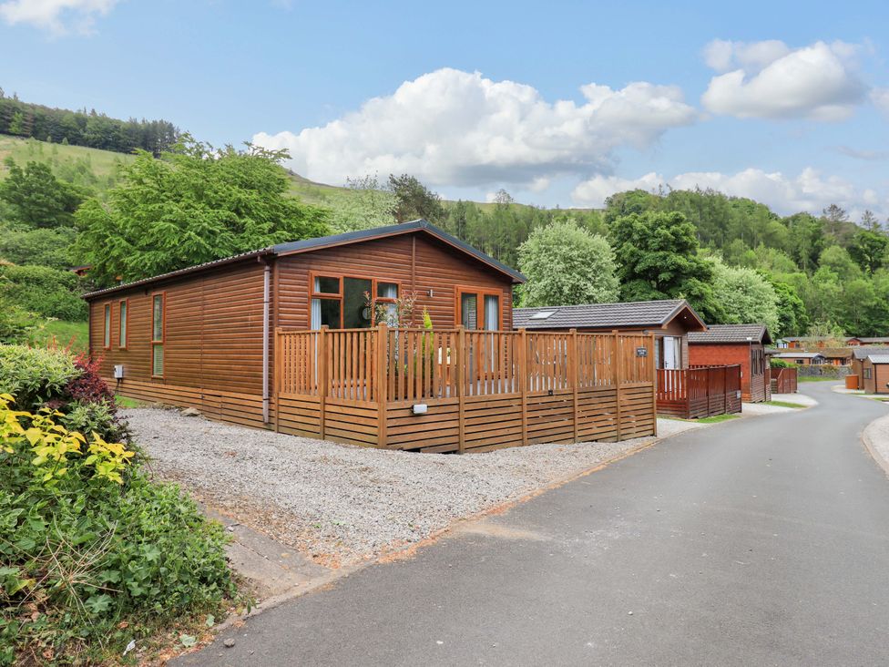 A wooden lodge with a fenced deck next to a paved road with other wooden buildings and trees in the background at Hollytree Lodge Beckside 1 Limefitt Park near Windermere