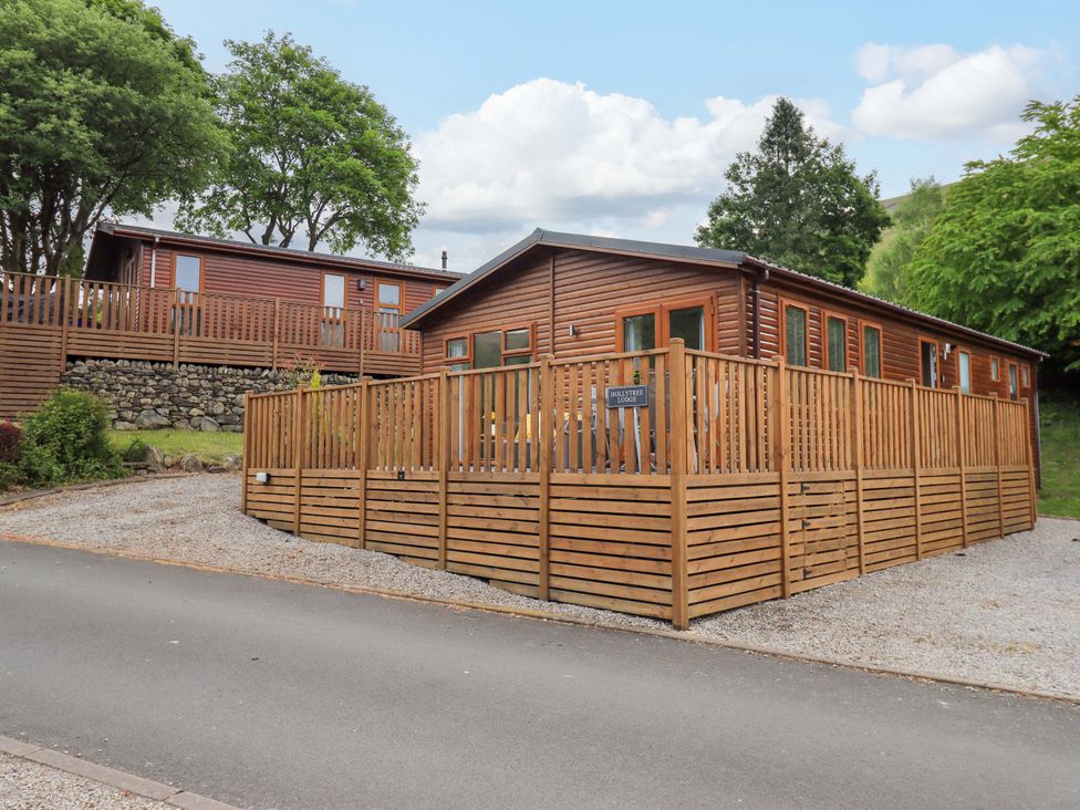 A wooden lodge with a fenced deck and gravel driveway surrounded by trees at Hollytree Lodge Beckside 1 Limefitt Park near Windermere